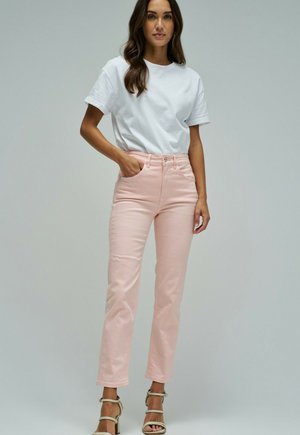 Mujer con pelo largo y oscuro que lleva una camisa blanca de manga corta, vaqueros rosa claro y sandalias beige de tacón, de pie con una mano en el bolsillo.
