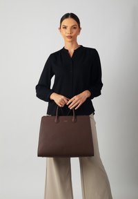 Brown leather tote bag with a structured design, dual rounded handles, and a textured surface. Features a zipper closure and subtle branding.
