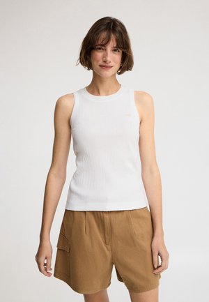 Femme aux cheveux bruns courts portant un haut blanc sans manches et un short marron ample, debout devant un fond clair uni.