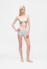 Kvinde står barfodet iført grå bh med blonder og højtaljede shorts mod en ensartet hvid baggrund.