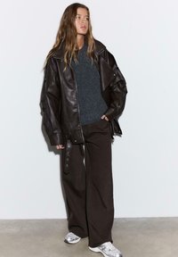 Hnedá kožená oversize bunda so širokým golierom, pod ňou sivý textúrovaný sveter, tmavohnedé nohavice a strieborné tenisky. Biele pozadie.