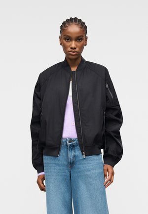 Femme aux cheveux tressés portant une veste bomber noire, un haut violet clair et un jean taille haute bleu, se tenant devant un fond uni.