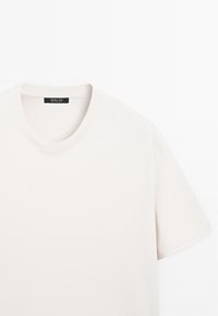 Jasnobeżowa bawełniana koszulka z okrągłym dekoltem i krótkimi rękawami. Wewnątrz kołnierza znajduje się metka. Gładka faktura, minimalistyczny design.