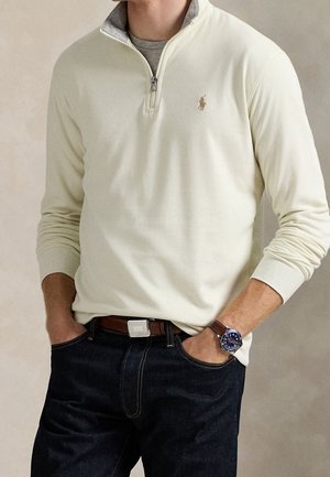Mand iført cremefarvet quarter-zip sweater, mørke jeans, brun læderbælte og blå armbåndsur med hænderne i lommerne.