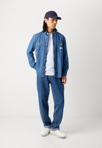 Blue denim shirt with buttons, white t-shirt underneath, blue denim pants, and white sneakers. Dark cap completes the outfit.