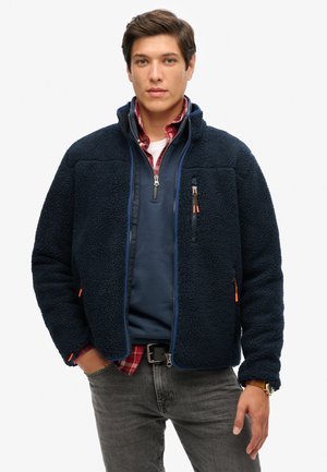 Navy fleece jacket with a high collar, zippered chest pocket, and orange zipper accents. Worn over a gray top and layered with a plaid shirt.