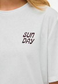 T-shirt bianco in cotone con una grafica testurizzata di colore marrone scuro che recita "SUN DAY" in un font giocoso e ondulato sulla parte sinistra.