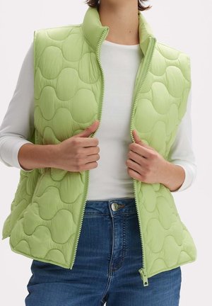 Gilet matelassé vert avec un motif de vagues, col montant et fermeture éclair à l'avant. Porté sur un haut blanc et associé à un jean bleu.