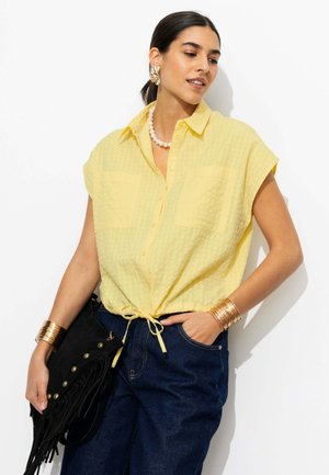 Femme portant une chemise jaune à manches courtes boutonnée et nouée à la taille, jean foncé, bracelets dorés, collier de perles et tenant un sac noir à franges.