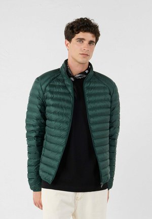 Hombre con una chaqueta acolchada verde oscuro sobre una camisa negra y pantalones blancos, de pie frente a un fondo claro y liso.