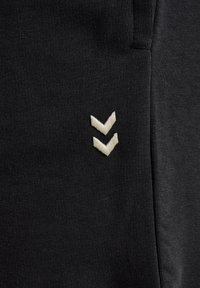 Schwarzer Stoff mit strukturiertem Strickmuster, der zwei kleine weiße, nach unten zeigende, bestickte Chevron-Zeichen nahe der Naht zeigt.