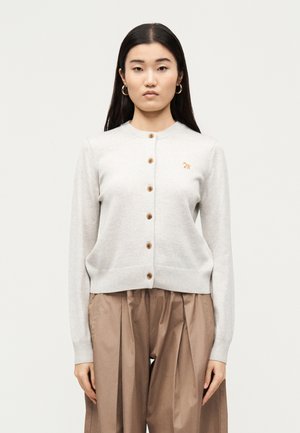 Žena oblečená v béžovom zapínanom kardigáne s malým vyšívaným zvieratkom a voľných hnedých plisovaných nohaviciach, stojaca pred jednoduchým svetlým pozadím.