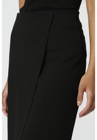 Jupe crayon noire avec un pli sur le côté, en tissu lisse. Elle présente une coupe ajustée et une silhouette épurée. Une main est posée sur le tissu.