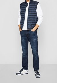 Granatowa pikowana kamizelka na zamek, biała koszula z długim rękawem, ciemne dżinsy oraz czarno-białe trampki. Tkanina o fakturze i swobodny krój.