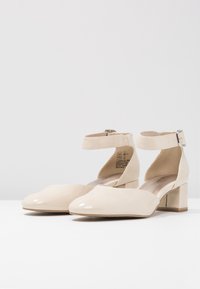 Béžové lakované lodičky se čtveratou špičkou, páskem kolem kotníku a masivním blokovým podpatkem. Hladký povrch s lesklým finišem.