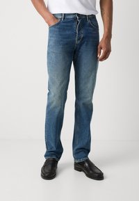 Jeans de mezclilla azul con corte recto, cinco bolsillos y un ligero desgaste. Combinados con mocasines negros sobre un fondo simple.
