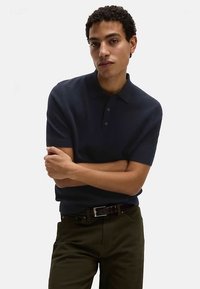 Polo blu navy in tessuto strutturato, maniche corte e colletto classico, abbinata a pantaloni verde oliva e cintura nera.