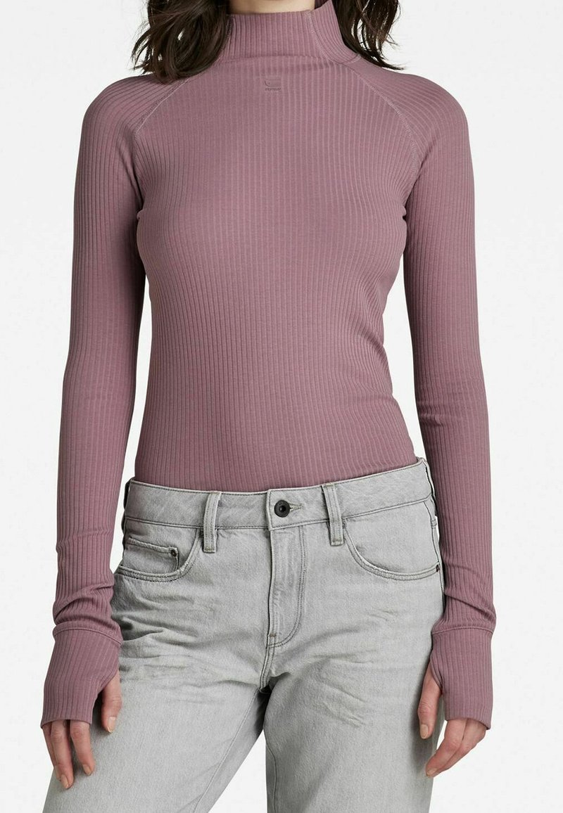 Femme portant un col roulé côtelé ajusté rose poussiéreux à manches longues avec des ouvertures pour les pouces, associé à un jean taille haute gris clair.