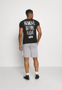 T-shirt noir avec le texte blanc "ENLEVER AVANT LE VOL" dans le dos, associé à un short gris et des baskets noires. Manches courtes, coupe régulière.