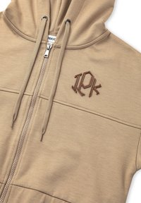 Beige rits hoodie met een capuchon, voorzien van ton-sur-ton stiksels en een geborduurd logo. Glad textuur en zilveren rits hardware.