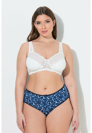 Frau trägt weißen Spitzen-BH und blaue Unterwäsche mit Leopardenmuster, steht vor hellem, einfarbigem Hintergrund und schaut nach vorn.