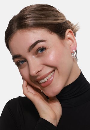 Orecchini in argento con un design elegante e curvato. La superficie è liscia, presentando un aspetto moderno e minimalista. Indossati da un modello sorridente.