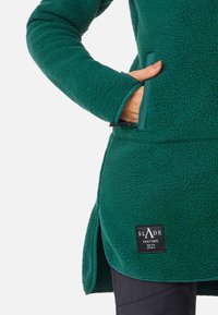 Cappotto in pile verde con tessuto testurizzato, tasche laterali e etichetta nera del marchio. Presenta un orlo curvo e un impatto di colore contrastante lungo i bordi.