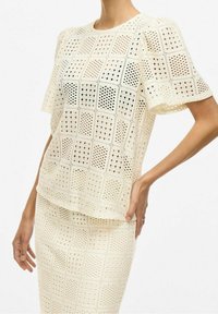 Kvinna som bär ett krämfärgat virkat set med topp och kjol med fyrkantiga mönster, en hand på höften och den andra avslappnad.