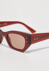 Ochelari de soare roșii din plastic cu ramă geometrică, lentile maronii și logo-ul argintiu "Ray-Ban" pe farmec. Detalii texturate pe brațe.