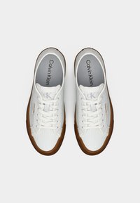 Baskets blanches Calvin Klein avec dessus texturé, lacets blancs et semelles en caoutchouc marron, vues de dessus sur un fond blanc.