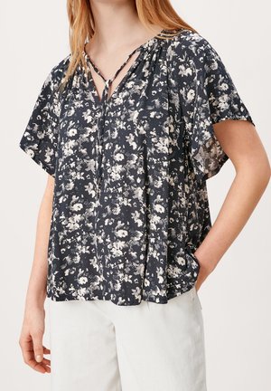 Mujer con una blusa floral negra y blanca suelta con mangas cortas y un lazo en el escote, combinada con pantalones blancos.