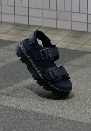 Melna ādas sandale ar divām regulējamām sprādzēm un biezām rupjām zolēm, novietota virs teksturēta bruģēta seguma un flīzētas sienas fona.