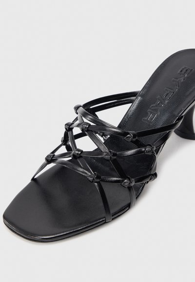 Schwarze hochhackige Sandalen mit geflochtenem Riemendesign, quadratischer Zehenform und glatter Leder-Oberfläche. Mit geknoteten Details für zusätzlichen Komplexität.