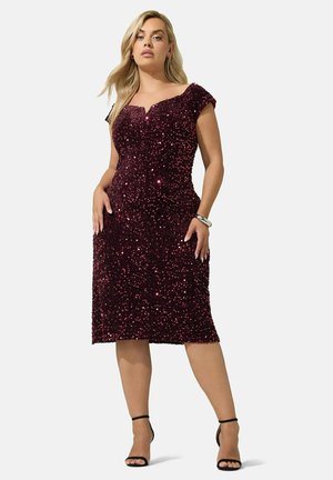 Femme portant une robe sans manches, longueur genoux, en sequins violet foncé, et des talons hauts noirs à brides, posant avec les mains près des hanches.