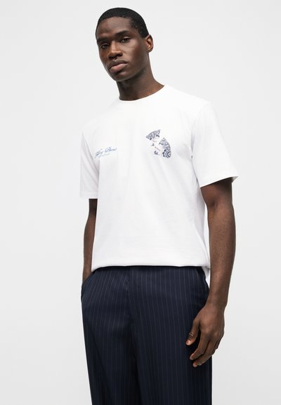 Άνδρας που φοράει λευκό γραφικό t-shirt και μπλε σκούρο παντελόνι με ρίγες, στέκεται με τα χέρια στις τσέπες μπροστά σε λευκό φόντο.