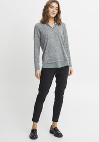 Harmaa neulepaita, jossa on V-pääntie, vihreä ruudullisen paidan helman päällä. Yhdistetty mustiin slim-fit-housuihin ja mustiin loafer-kengiin.
