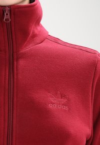 Červená bunda na zip z měkkého materiálu, s vysokým límcem, jemně vyraženým logem Adidas na hrudi a žebrovanými detaily na ramenou.