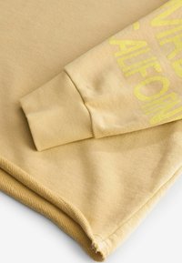 Manga de sudadera beige con puño acanalado y texto amarillo parcialmente visible, sobre un fondo de tela beige a juego.
