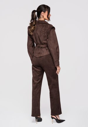 Kvinde med langt krøllet hestehale iført en brun jakke med bælte og matchende bukser, stående med ryggen til, i sorte højhælede sko.