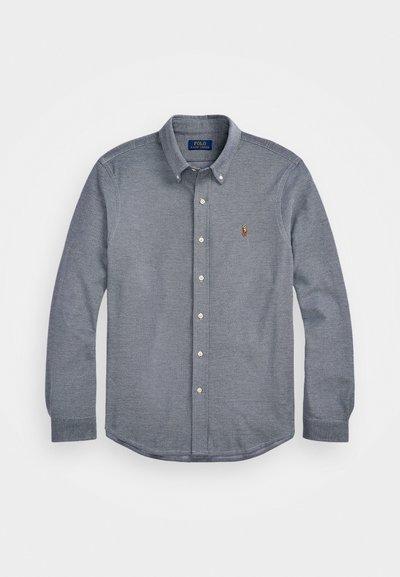 Knappskjorte i mørk blått stoff med teksturert mønster. Har spredt krage, lange ermer og en liten brodert logo på brystet.