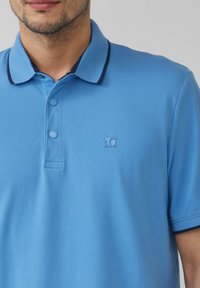 Lichtblauwe polo met marineblauwe kraagafwerking, twee-knopsluiting en klein logo op de borst. Textuurstof met een casual pasvorm.