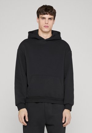 Young man wearing plain black hooded sweatshirt and matching black pants, standing against a light gray background.