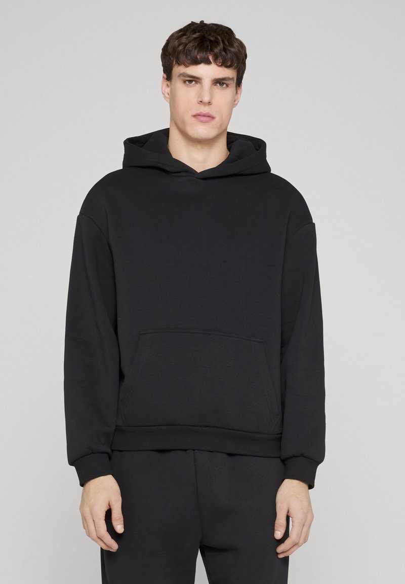 Mlad moški nosi enostavno črno jopico s kapuco in primerne črne hlače, stoji na svetlo sivi podlagi.