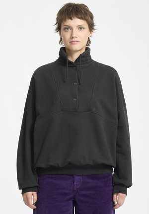 Sweat à col montant noir avec patte de boutonnage, ainsi que des poignets et un ourlet côtelés. Présente un logo minimaliste sur la poitrine et un tissu texturé.