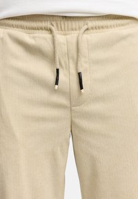 Beige ribbed sweatpants with an elastic waistband, drawstring with black accents, and side pockets. Textured fabric adds visual interest.