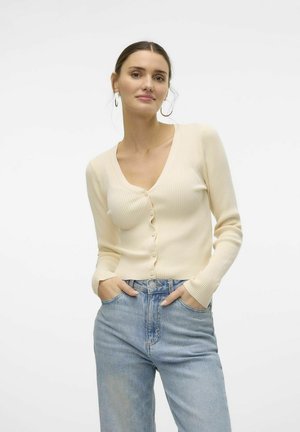 Cardigan crème côtelé avec un col en V, doté d'une fermeture par boutons et de manches longues. Le modèle porte un jean taille haute bleu clair, les mains dans les poches.