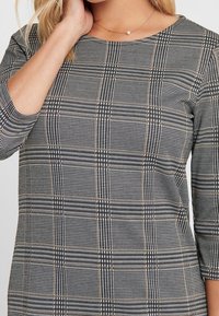 Chemisier à carreaux noir et blanc avec des manches trois-quarts, un col rond et de subtils accents marron dans le motif en tartan. Texture lisse.