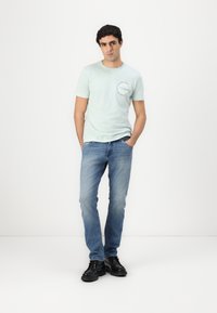 T-shirt verde menta a manica corta con scollatura rotonda e stampa sul petto, abbinata a jeans denim blu chiaro e scarpe nere.
