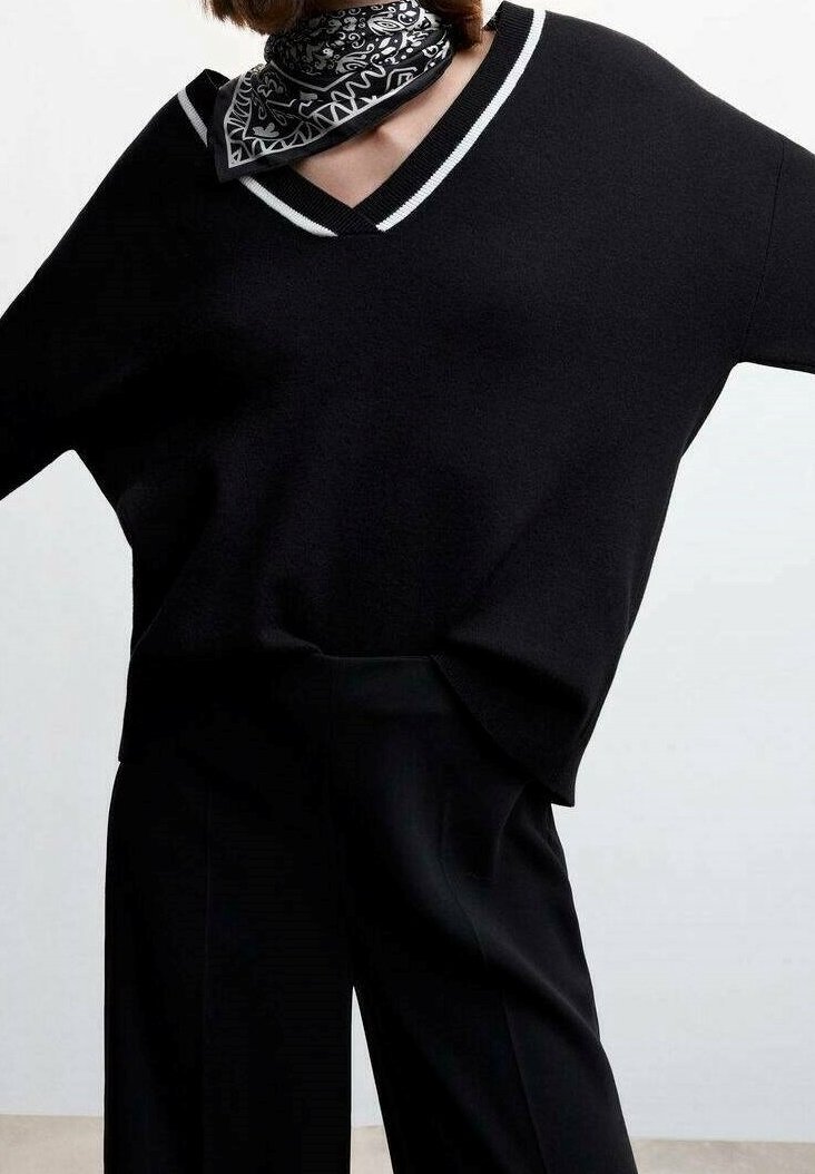 Personne portant un pull noir en V avec une bordure blanche, un pantalon noir et un foulard noir et blanc à motif, posant devant un fond uni.