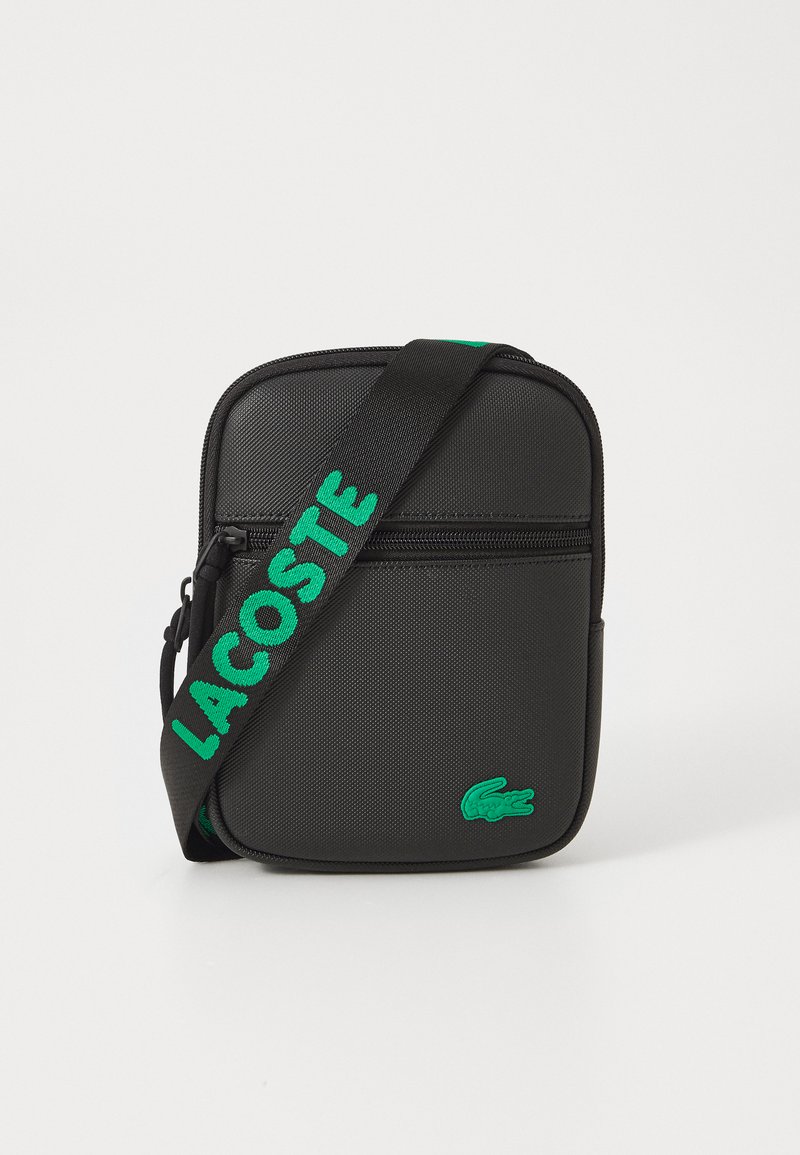 Borsa a tracolla nera con tessuto testurizzato, un logo verde e la scritta "LACOSTE" stampata sulla tracolla regolabile; presenta un compartimento principale con zip.